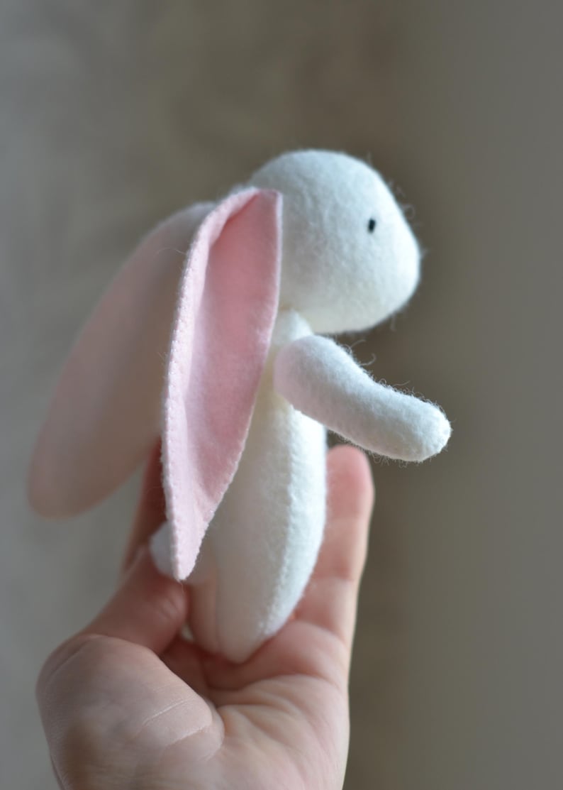May include: A handmade white stuffed bunny with pink inner ears. The plush toy has a small black eye and is held in a hand. The bunny is made of soft fabric and is a cute, whimsical design.