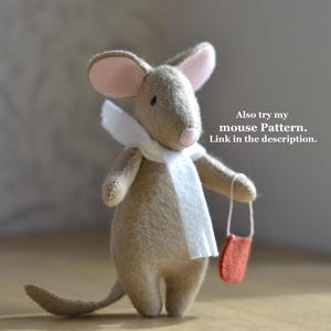 May include: A handmade felt mouse with a white scarf and a small red bag. The mouse has pink ears and a long tail. The text "Also try my mouse Pattern. Link in the description." is visible.