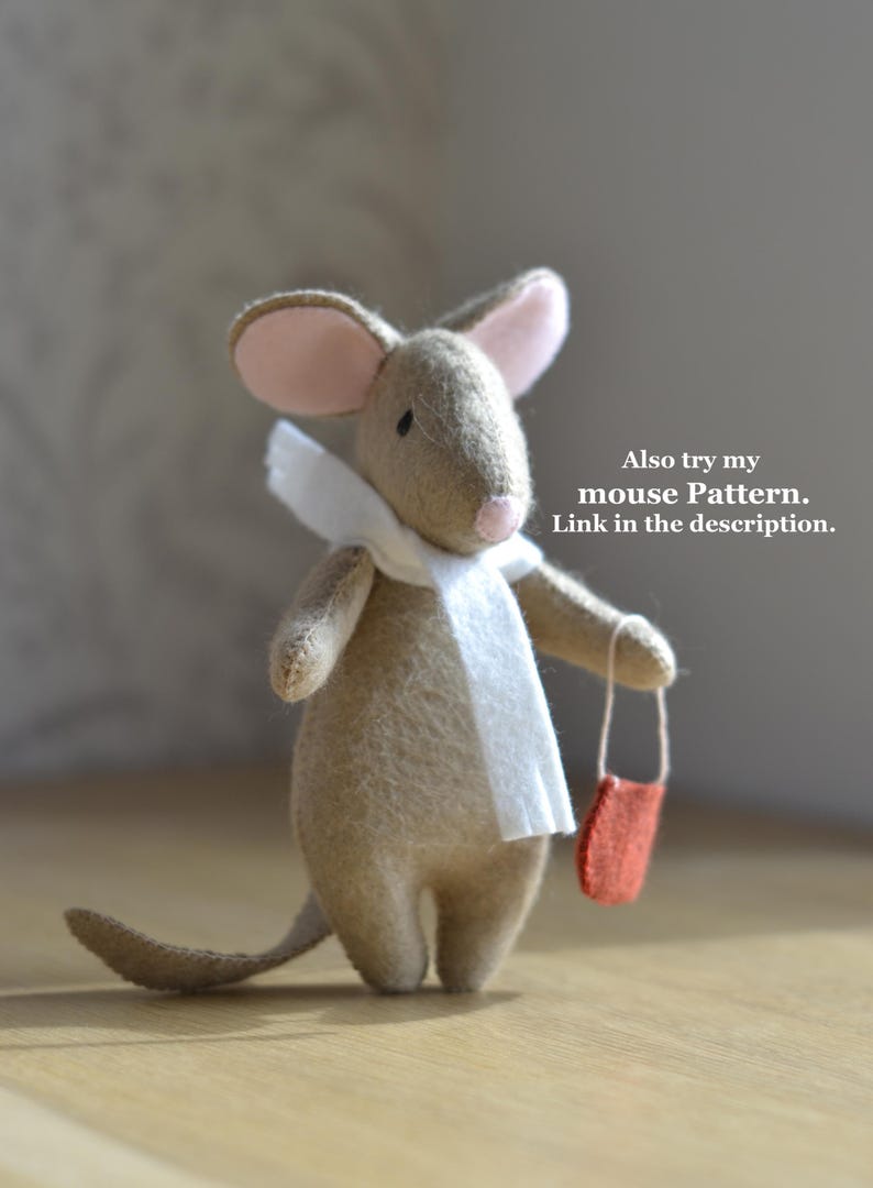 May include: A handmade felt mouse with a white scarf and a small red bag. The mouse has pink ears and a long tail. The text "Also try my mouse Pattern. Link in the description." is visible.