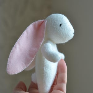 May include: A handmade white stuffed bunny with long, pink-lined ears. The plush toy has small black eyes and is held in a person's hand. The bunny is a soft, cuddly toy.