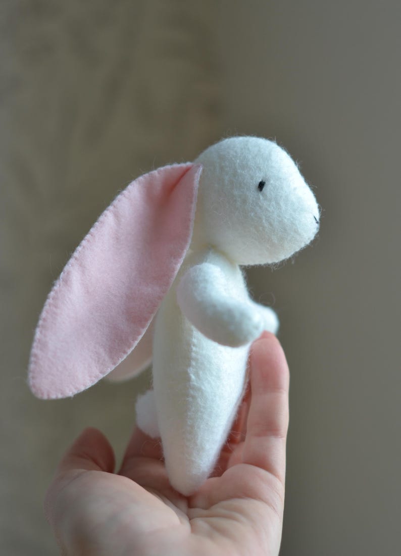 May include: A handmade white stuffed bunny with long, pink-lined ears. The plush toy has small black eyes and is held in a person's hand. The bunny is a soft, cuddly toy.