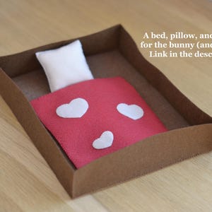 May include: A miniature bed set for a bunny or mouse, featuring a brown cardboard bed frame, a white pillow, and a red blanket adorned with white heart shapes. The text on the image reads: "A bed, pillow, and blanket for the bunny (and mouse). Link in the description."