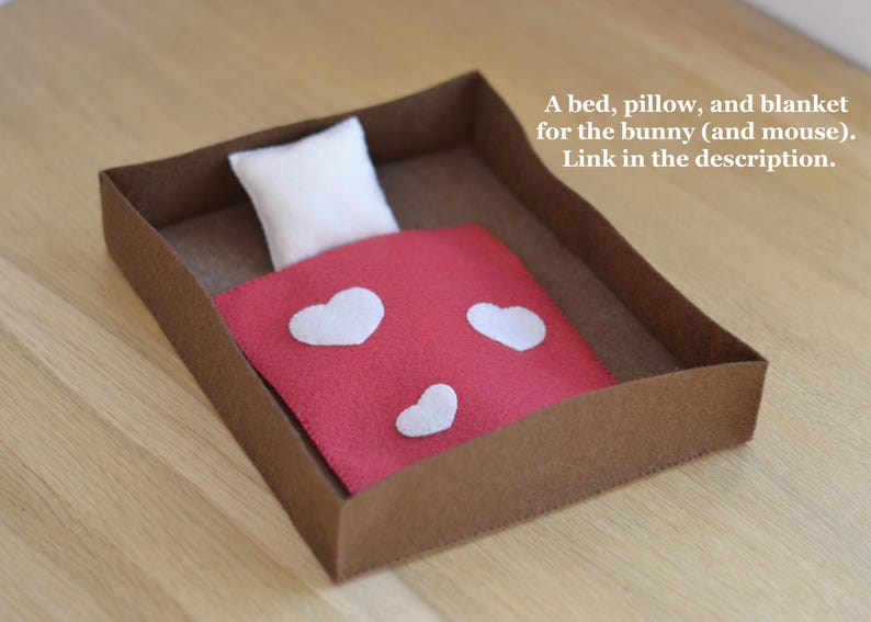 May include: A miniature bed set for a bunny or mouse, featuring a brown cardboard bed frame, a white pillow, and a red blanket adorned with white heart shapes. The text on the image reads: "A bed, pillow, and blanket for the bunny (and mouse). Link in the description."