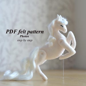 Könnte beinhalten: Ein weißes Einhorn-Spielzeug aus Filz mit fließender Mähne und Schweif, das sich aufbäumt. Der Text "PDF felt pattern Photos step by step" ist im Bild zu sehen.