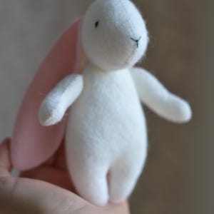 May include: A small, handmade white bunny plush toy with pink inner ears. The bunny has black embroidered eyes and a stitched nose. The toy is held in a person's hand, showcasing its size and soft texture. A charming, handcrafted item.