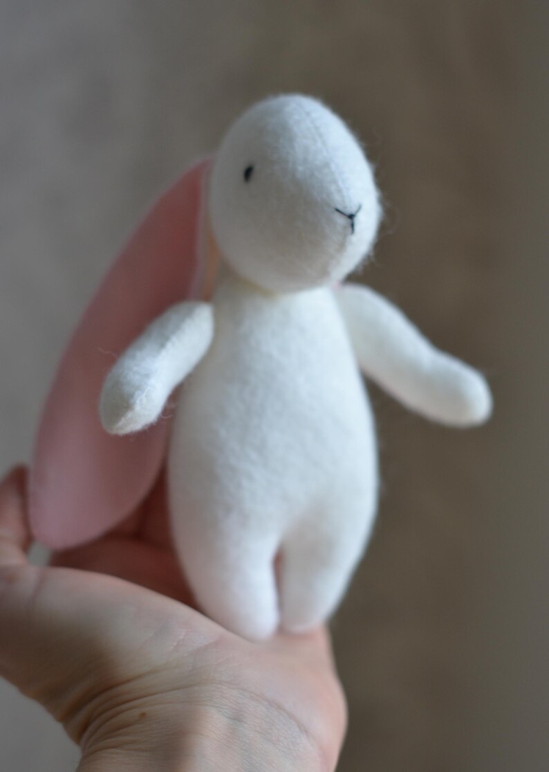 May include: A small, handmade white bunny plush toy with pink inner ears. The bunny has black embroidered eyes and a stitched nose. The toy is held in a person's hand, showcasing its size and soft texture. A charming, handcrafted item.