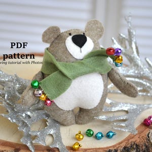 Puede incluir: Un oso de fieltro hecho a mano con una bufanda verde y una guirnalda de campanas de colores. El oso tiene una barriga blanca y una nariz negra. La imagen incluye el texto "PDF pattern sewing tutorial with Photos".