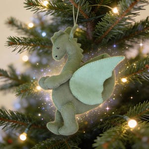 Op de afbeelding: Een handgemaakte, lichtgroene vilten draak kerstversiering met een lichtgroene vleugel. De draak hangt aan een gouden draad aan een kerstboom. De versiering heeft een grillig ontwerp en is versierd met kleine, zwarte kraaloogjes.