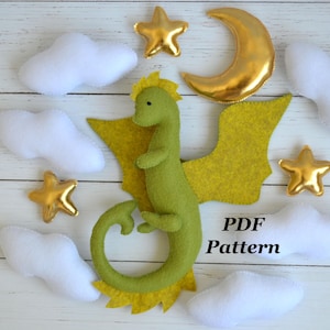 May include: A felt dragon with yellow wings and accents, surrounded by white clouds, gold stars, and a gold crescent moon. The text "PDF Pattern" is visible. This is a craft project.