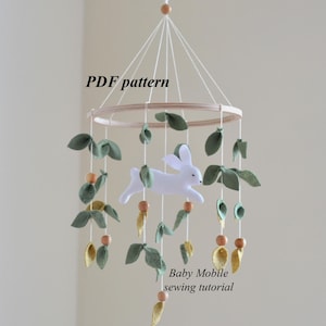 Può includere: Mobile per bambini fatto a mano con un coniglietto in feltro bianco, foglie verdi e decorazioni a forma di carota. Il mobile è sospeso a un cerchio di legno con perline di legno e spago. Il testo sull'immagine recita "PDF pattern" e "Baby Mobile sewing tutorial."