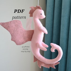 Peut inclure: Mobile en forme de dragon rose en feutre, avec des ailes et une queue enroulée. Le texte "PDF pattern" est au-dessus du dragon, et "45 photos sewing step by step" est en dessous.