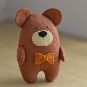 May include: A brown felt bear plush toy featuring a cream-colored muzzle, a small black nose, two tiny black eyes, and a bright orange bow tie attached to the chest. The bear has rounded ears and a simple, stitched body construction.