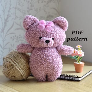 May include: A handmade pink teddy bear with a bow, black eyes, and a black nose. The bear is made of a textured material. A ball of twine and a small potted plant are next to the bear. The text "PDF pattern" is visible.