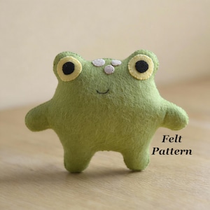 Puede incluir: Una rana de fieltro verde hecha a mano con grandes ojos amarillos y negros, pequeños puntos blancos y una boca sonriente. La rana tiene los brazos y las piernas extendidos. El texto a la derecha dice "Felt Pattern".