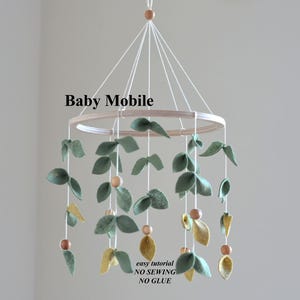 May include: A baby mobile with a wooden ring, from which hang felt leaves in green and yellow, and wooden beads. The mobile is suspended by white strings. Text on the image reads "Baby Mobile" and "easy tutorial NO SEWING NO GLUE".