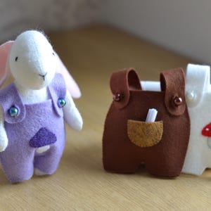 May include: A white felt bunny wearing purple overalls with iridescent buttons. Next to the bunny are two pairs of felt overalls, one brown and one white, with a mushroom applique. The overalls have small pockets and button details.