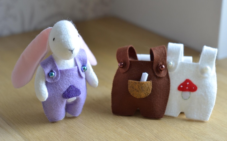 May include: A white felt bunny wearing purple overalls with iridescent buttons. Next to the bunny are two pairs of felt overalls, one brown and one white, with a mushroom applique. The overalls have small pockets and button details.