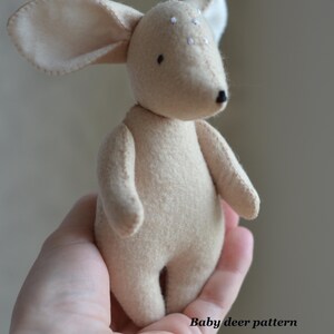 May include: A handmade beige deer plush toy with large ears and a small black nose. The deer has white spots on its head and is held in a hand. Text on the image reads "Baby deer pattern" and "Link in the description".