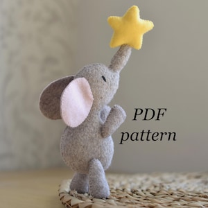 Könnte beinhalten: Ein graues Elefanten-Spielzeug aus Filz, das mit seinem Rüssel nach einem gelben Stern greift. Der Elefant hat rosa Innenohren und steht auf einer gewebten Oberfläche. Der Text rechts lautet "PDF pattern".
