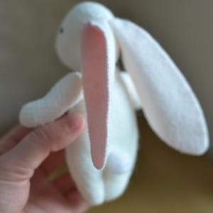May include: A white stuffed bunny with long ears and a small, fluffy tail. The inside of the ears is a soft pink. The bunny is made of a soft material and is held by a hand.