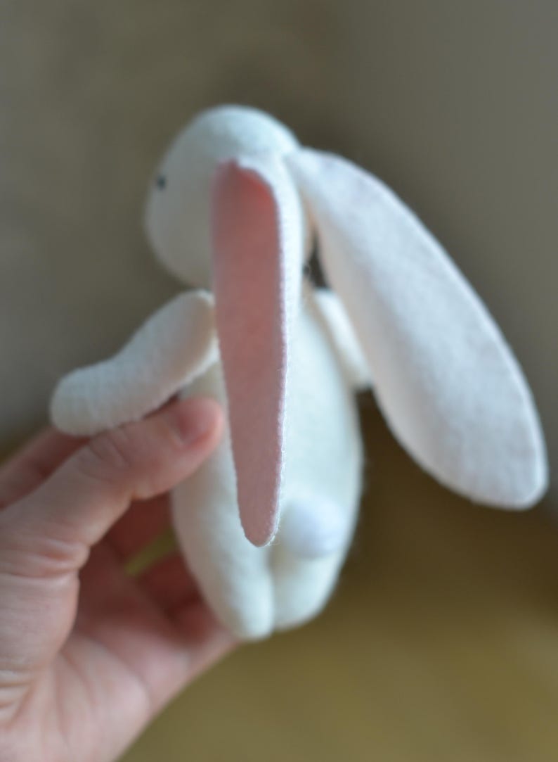 May include: A white stuffed bunny with long ears and a small, fluffy tail. The inside of the ears is a soft pink. The bunny is made of a soft material and is held by a hand.