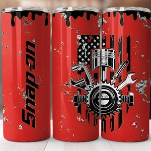 May include: Red tumbler with a black and white design featuring tools, a piston, and the American flag. The word "Snap-on" is printed vertically in black on the side of the tumbler. The tumbler has a black lid.
