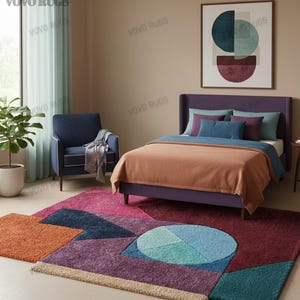 May include: A bedroom scene featuring a geometric patterned rug in shades of orange, blue, purple, and teal. A bed with a purple headboard and a peach-colored blanket is in the background. A blue armchair and a framed abstract art piece complete the room.