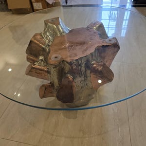 May include: A round coffee table with a clear glass top and a base crafted from a metallic, multi-faceted tree stump sculpture. The base features a combination of polished copper and silver-toned metal surfaces with an irregular, organic shape.