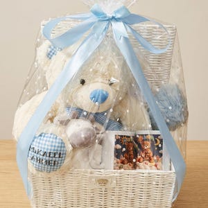 May include: A white wicker gift basket wrapped in clear cellophane and tied with a light blue ribbon. The basket contains a teddy bear, a mug with a teddy bear design, and other baby items. The teddy bear has a blue nose and a blue and white checkered patch.