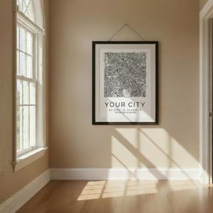 Op de afbeelding: Een zwart-wit stadsplattegrond in een zwarte lijst hangt aan een beige muur. De print toont een gedetailleerde kaart met de tekst "YOUR CITY" en coördinaten. De kamer heeft een houten vloer en een groot boogvormig raam.