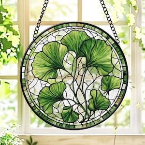 May include: Round stained glass window with a black metal frame and chain. The design features green ginkgo leaves with intricate details, set against a white and light green background. The window is hanging in front of a window.