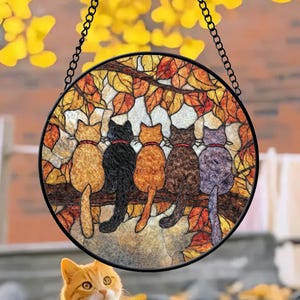 Puede incluir: Una vidriera redonda con cinco gatos de varios colores sentados en una rama. Los gatos están de espaldas al espectador, cada uno con un collar rojo o negro. El fondo incluye hojas de otoño y una cadena negra para colgar.