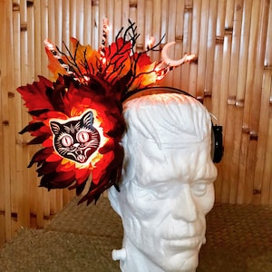 Puede incluir: Una diadema de Halloween con una cara de gato negro y hojas rojas y naranjas. La diadema está iluminada con luces blancas y tiene una luna creciente en el lateral.