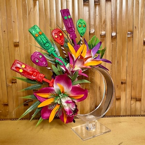 Puede incluir: Un arreglo floral colorido con una variedad de flores tropicales y hojas verdes. El arreglo se muestra en un soporte de plástico transparente. Las flores están acentuadas con figuras de estilo tiki de colores.
