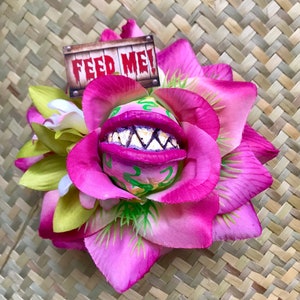 Puede incluir: Una flor rosa con una boca verde y morada que dice "¡Alimentame!" en un letrero de madera. La flor está hecha de tela y tiene un aspecto realista.