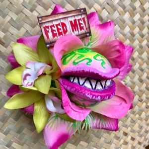 Puede incluir: Una flor rosa y verde con una boca grande y dentada y las palabras "FEED ME!" en un letrero de madera. La flor está hecha de tela y es una decoración divertida y caprichosa.
