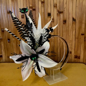 Puede incluir: Una diadema negra con un lirio blanco, un helecho negro y un gato negro con ojos verdes. La diadema está sobre un soporte de acrílico transparente.