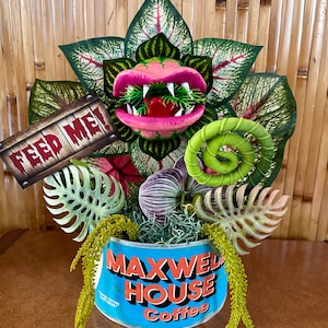 May include: A pink and green carnivorous plant with a mouth that says "Feed Me!" in red letters. The plant is in a blue and white Maxwell House coffee tin.
