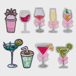 May include: A collection of embroidered patches featuring various cocktail and drink designs. The patches include a pink milkshake, wine glasses, champagne flutes, and margarita glasses, each with a decorative bow. The patches have a silver border.