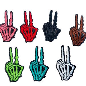 May include: A collection of seven embroidered patches in the shape of skeleton hands making the peace sign. The patches are in various colors, including pink, red, black, brown, green, teal, and white. Each patch has a glittery border.