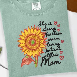 May include: A sage green t-shirt featuring a sunflower graphic with the words "She is strong, fearless, warm, loving, patient, selfless Mom" in black script, accented with red hearts.