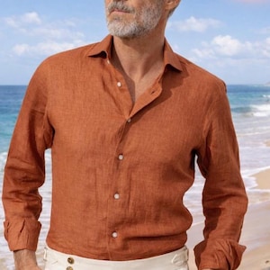 May include: A burnt orange linen shirt with long sleeves and a button-down front. Paired with cream-colored pleated trousers. The outfit is worn on a beach with blue water and a sandy shore in the background.