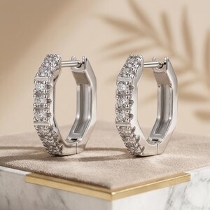May include: A pair of silver-toned hoop earrings with a hexagonal shape. Each earring features a row of small, clear, round-cut stones embedded along the front-facing edge. The earrings have a hinged post mechanism for secure closure.