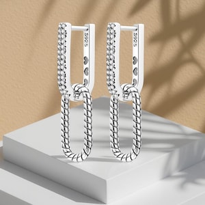 Puede incluir: Un par de pendientes de plata. La sección superior es rectangular y está engastada con pequeñas piedras transparentes. La sección inferior tiene forma ovalada con un diseño texturizado en forma de cuerda. Los pendientes están conectados por un lazo texturizado. El texto "S925" es visible.