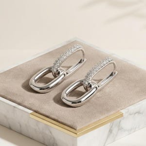 May include: A pair of silver-toned hoop earrings featuring a double-link design. The upper portion of each earring is an elongated oval encrusted with small, clear, sparkling stones, while the lower portion is a smooth, polished silver-toned oval link.
