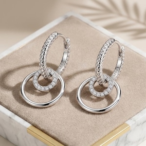 May include: A pair of silver hoop earrings featuring a twisted rope texture on the upper hoop and a smaller, circular charm encrusted with clear, sparkling stones suspended from each. The earrings are stamped with S925 to indicate sterling silver.