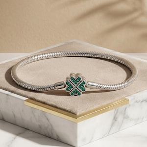 Könnte beinhalten: Ein silberfarbenes Schlangenkettenarmband mit einem zentralen Verschluss in Form eines vierblättrigen Kleeblatts. Das Kleeblatt ist mit kleinen, leuchtend grünen Steinen besetzt, die in jedes der vier herzförmigen Blätter eingelassen sind.