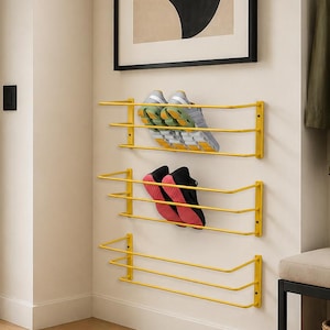 May include: Three yellow metal wall-mounted shoe racks with horizontal bars. The top rack holds a pair of grey and yellow athletic trainers, and the middle rack holds a pair of black and pink athletic trainers. The bottom rack is empty.