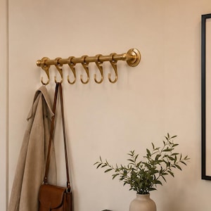 May include: A gold-colored metal coat rack with six hooks is mounted on a cream-colored wall. A tan coat and a brown leather purse hang from the hooks. A small vase with greenery sits on a black cabinet.