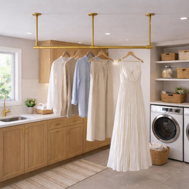 Pu&ograve; includere: Un appendiabiti in metallo dorato montato a soffitto e sospeso tramite tre aste verticali. L'appendiabiti sostiene diversi capi su grucce in legno, tra cui una camicia bianca, una camicia beige, una camicia a righe azzurre, una gonna beige e un abito bianco.